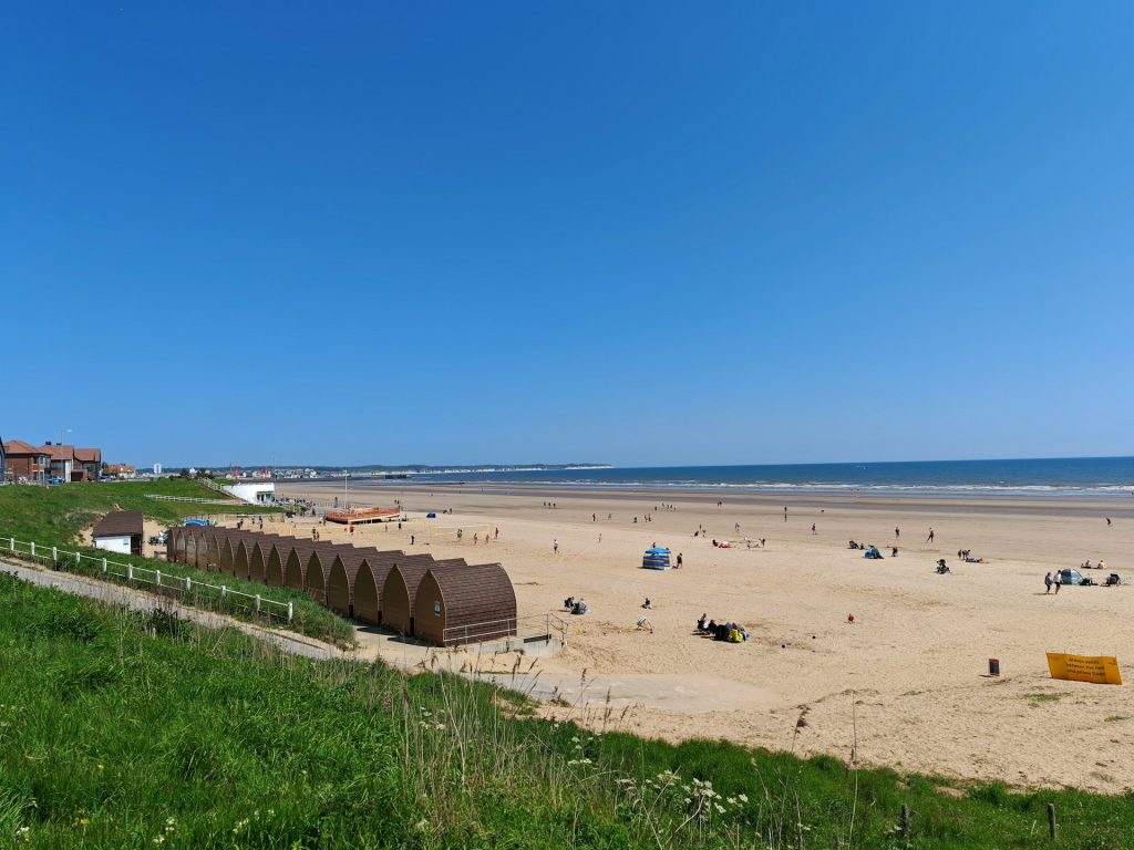 Panoramic view of beach huts and people on Bridlington beach under a cloudless, blue sky.