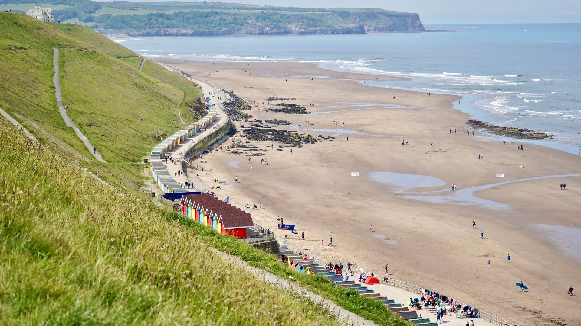 A side view of Whitby Beach featuring grassy hills and colourful beach huts on a clear day.