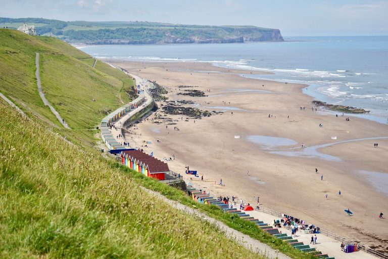 A side view of Whitby Beach featuring grassy hills and colourful beach huts on a clear day.