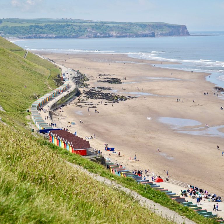 A side view of Whitby Beach featuring grassy hills and colourful beach huts on a clear day.