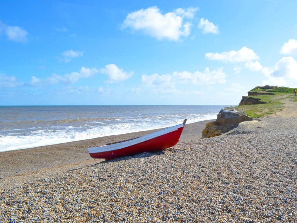 Red dinghy sitting on the shingle beach at Weybourne under a blue sky.